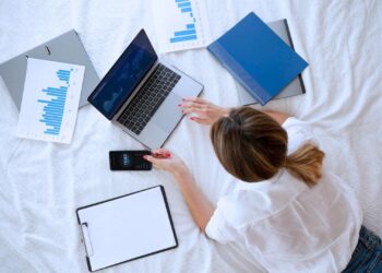 Mulher deitada na cama manuseando um notebook. Ela está rodeada de anotações.