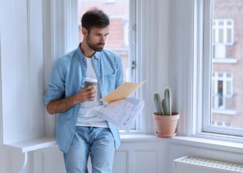 Homem em pé analisando alguns papéis enquanto segura um copo de café.