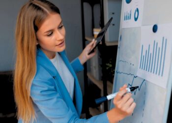 Mulher de terno azul escrevendo em uma lousa branca com alguns gráficos.