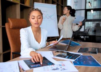 Mulher analisando dados e indicadores de uma empresa. Ela está rodeada de papéis e um notebook sobre a mesa.
