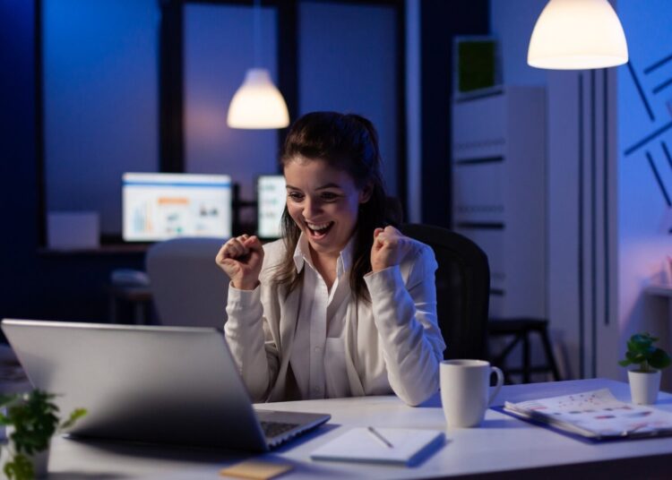 Mulher vibrando olhando para o notebook. Ela está em um escritório.