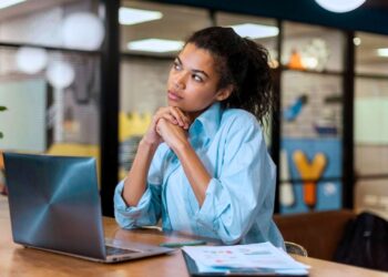 Mulher pensativa apoiada sobre uma mesa de escritório. Ao lado dela há um notebook e alguns papéis.