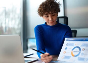Mulher trabalhando em um escritório. Ela está usando um casaco azul e analisando alguns documentos.