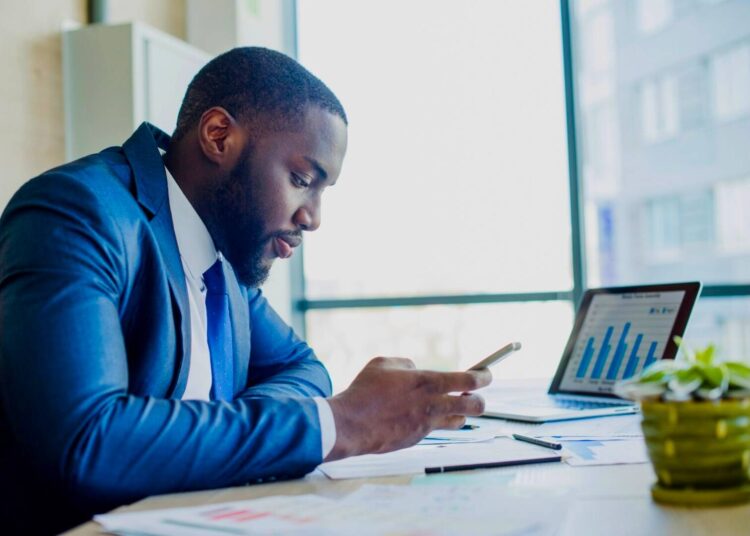 Homem olhando para o celular. Ele está apoiado sobre uma mesa onde há um notebook e vários papéis.