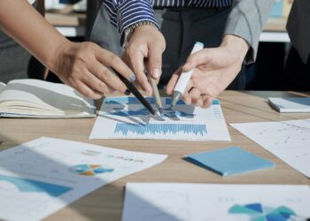 Mãos segurando canetas e apontando para papéis com vários dados e gráficos.