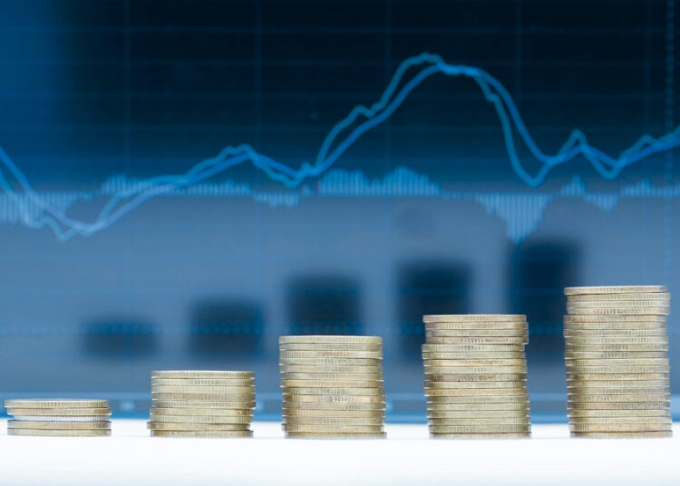 Cinco montes de moedas empilhadas. Atrás delas há um gráfico azul com tendência de crescimento.