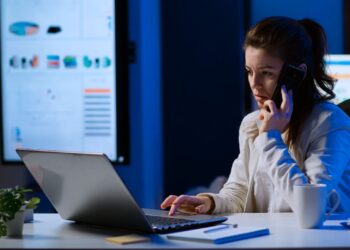Mulher em ligação no celular enquanto consulta informações no notebook.
