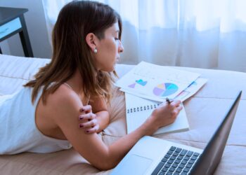 Mulher deitada na cama enquanto anota em um planner. Ao seu lado há um notebook.