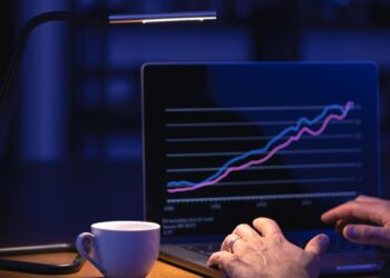 Homem manuseando um notebook. Na tela há um gráfico com tendência de alta e ao seu lado uma xícara.