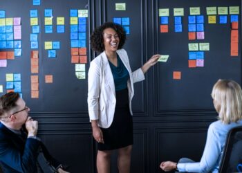 Reunião de trabalho. Há uma mulher sorrindo enquanto cola post-its na parede.