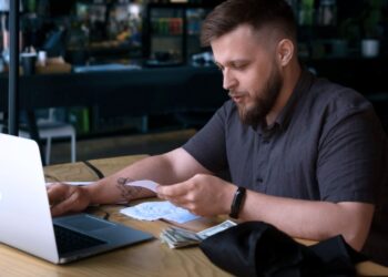 Homem mexendo no notebook enquanto analisa notas fiscais.