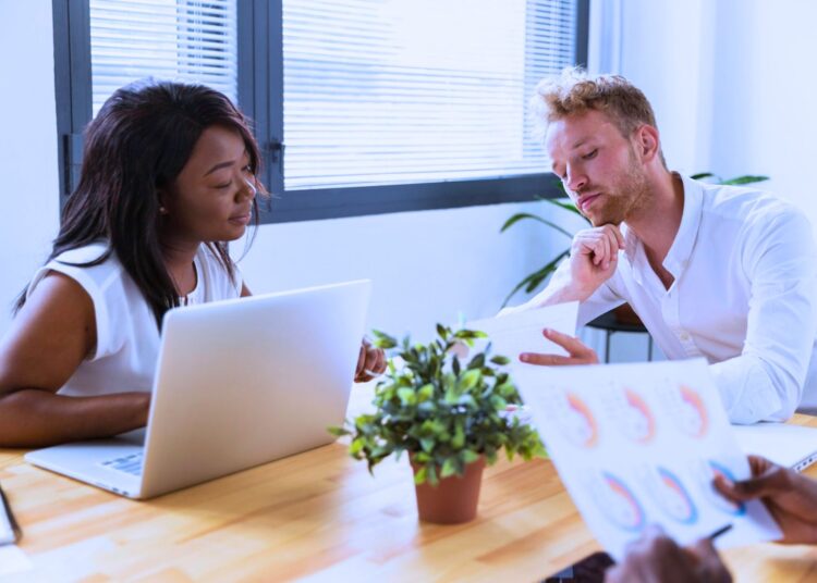 Uma mulher e um homem conversando sobre finanças. Ela está com um notebook enquanto ele mostra um papel com informações.