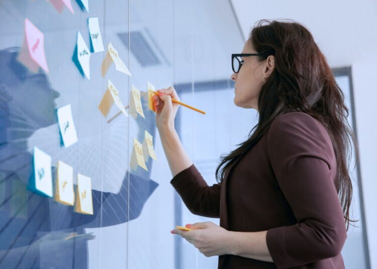 Mulher preenchendo post-its colados em uma parede de um escritório.