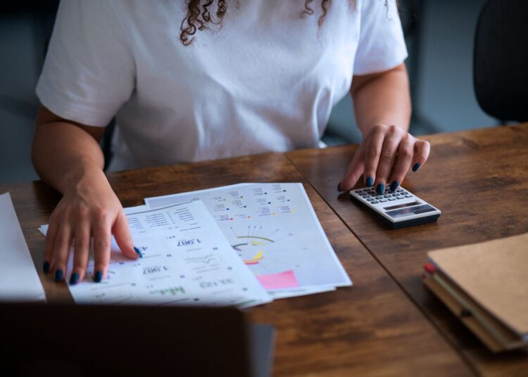 Mulher analisando dados financeiros da empresa. Ela está fazendo contas em uma calculadora.