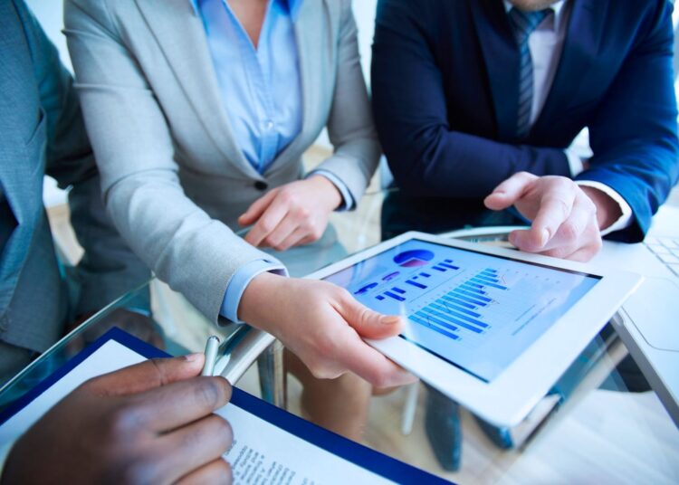 Três executivos conversando ao redor da mesa. Eles observam o tablet que está na mão de um deles.