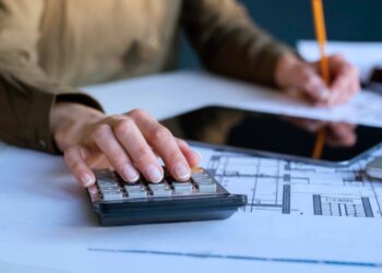 Mulher escrevendo em um papel sobre a mesa. Ao seu redor há uma calculadora e um tablet.