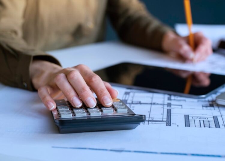 Mulher escrevendo em um papel sobre a mesa. Ao seu redor há uma calculadora e um tablet.