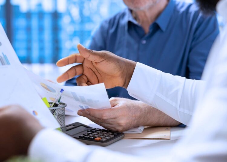 Dois homens analisando indicadores financeiros de uma empresa. Eles estão com papéis nas mãos e uma calculadora sobre a mesa.