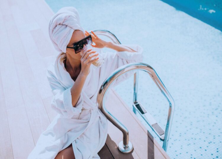 Mulher de roupão tomando um drink à beira da piscina.