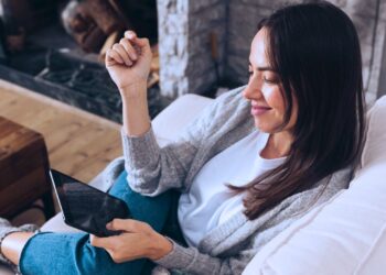 Mulher sorrindo sentada no sofá enquanto olha para o seu tablet.