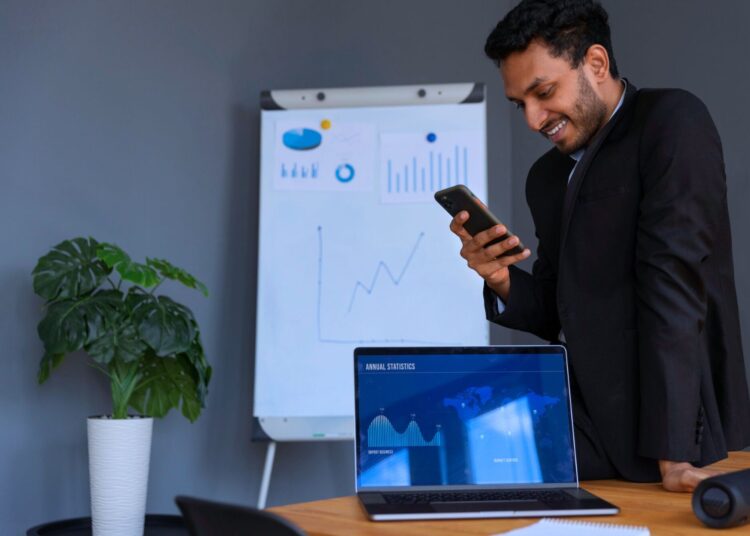 Homem de terno em um ambiente corporativo, sorrindo enquanto usa um smartphone, com gráficos exibidos em um notebook e em um quadro branco ao fundo.