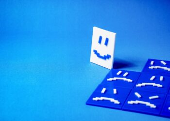 Imagem abstrata em tons de azul com blocos representando rostos em 3D. No canto, destaca-se um rosto branco sorridente, contrastando com rostos azuis tristes ao fundo.