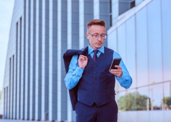 Homem com roupa social na rua carregando o terno e mexendo no celular.