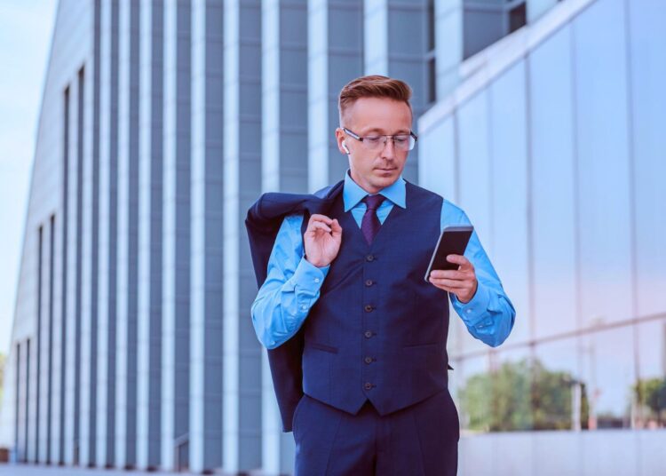 Homem com roupa social na rua carregando o terno e mexendo no celular.