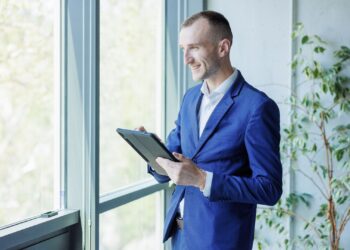 Homem com roupa social segurando um tablet olhando para a janela.