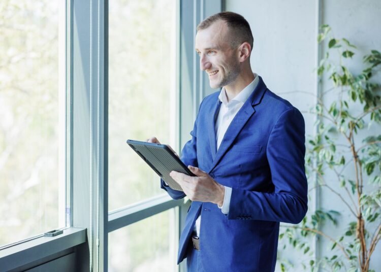 Homem com roupa social segurando um tablet olhando para a janela.
