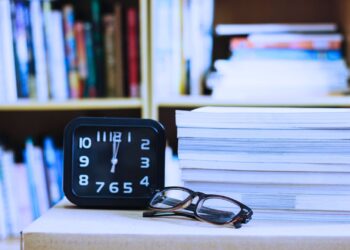 Pilha de livros ao lado de um relógio e um óculos.