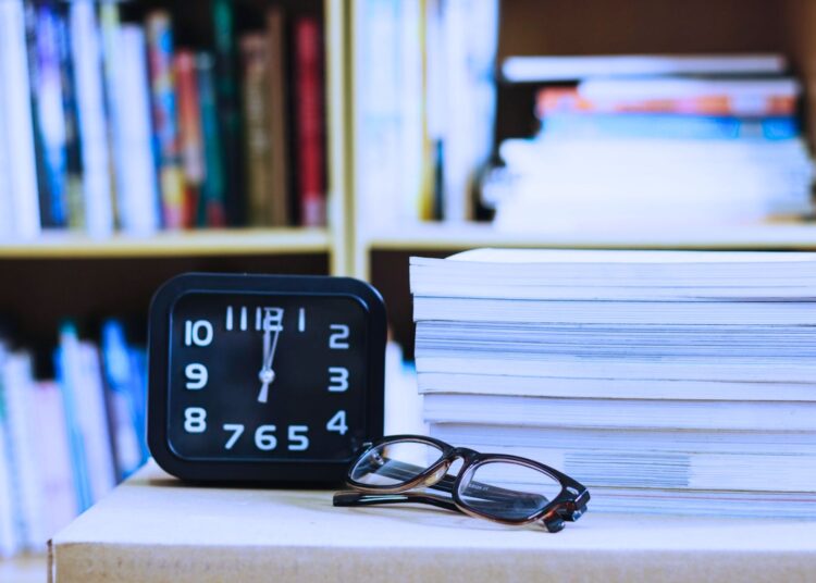 Pilha de livros ao lado de um relógio e um óculos.