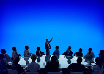 Imagem de uma reunião corporativa com pessoas sentadas ao redor de uma mesa em formato oval, enquanto um homem de pé faz uma apresentação em frente a um fundo azul brilhante.