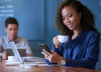Mulher tomando um café enquanto olha para o celular. Ela está em uma sala de reunião.