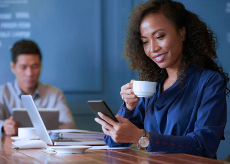 Mulher tomando um café enquanto olha para o celular. Ela está em uma sala de reunião.