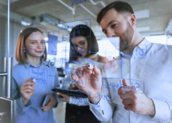 Equipe colaborando em um planejamento estratégico, escrevendo e discutindo ideias em um painel de vidro, representando táticas e organização para alcançar objetivos empresariais.