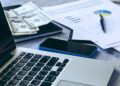 Imagem de uma mesa de trabalho com um laptop, notas de dólares, smartphone e gráficos financeiros impressos.