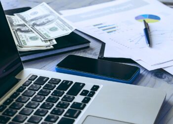 Imagem de uma mesa de trabalho com um laptop, notas de dólares, smartphone e gráficos financeiros impressos.