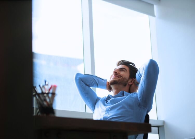 Homem sentado em uma cadeira de escritório, vestindo camisa azul, com as mãos atrás da cabeça e um sorriso no rosto, olhando para fora de uma grande janela.