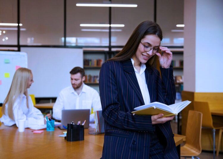 Profissional sorridente lendo um livro em um ambiente de trabalho colaborativo, com colegas ao fundo em discussão, simbolizando aprendizado contínuo e troca de conhecimentos.