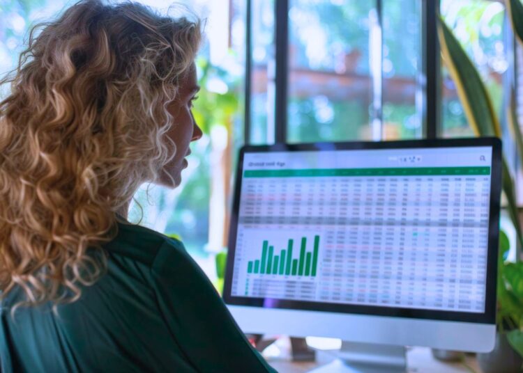 Profissional analisando gráficos e tabelas financeiras em uma tela de computador, em um ambiente de trabalho com luz natural e plantas ao fundo, representando análise de dados e planejamento estratégico.