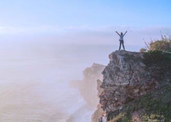 Pessoa comemorando no topo de um penhasco com os braços erguidos, representando automotivação, superação e conquista de objetivos em um cenário de natureza exuberante.