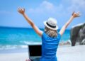 Pessoa trabalhando remotamente em uma praia, com um laptop e braços erguidos em celebração, simbolizando liberdade e equilíbrio entre trabalho e lazer.