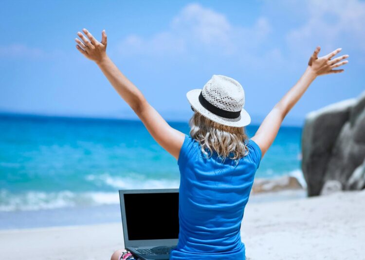 Pessoa trabalhando remotamente em uma praia, com um laptop e braços erguidos em celebração, simbolizando liberdade e equilíbrio entre trabalho e lazer.