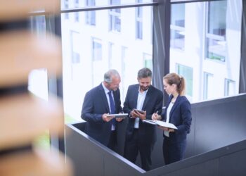Trio de profissionais em um ambiente corporativo discutindo estratégias e analisando dados em tablets e cadernos, representando colaboração e liderança no trabalho.