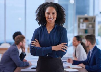Mulher confiante em posição de liderança em um ambiente de trabalho colaborativo, com uma equipe ao fundo engajada em discussões, simbolizando liderança democrática e inclusiva.