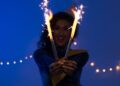 Mulher segurando sparkles acesos em um ambiente festivo e iluminado, simbolizando celebração e entusiasmo com foco em metas e resoluções para o Ano Novo.