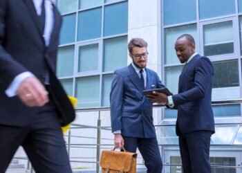 Dois profissionais discutindo documentos em um tablet em frente a um prédio corporativo, representando análise de patrimônio líquido e interação empresarial.