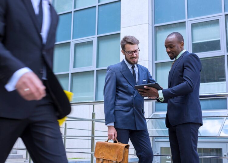 Dois profissionais discutindo documentos em um tablet em frente a um prédio corporativo, representando análise de patrimônio líquido e interação empresarial.