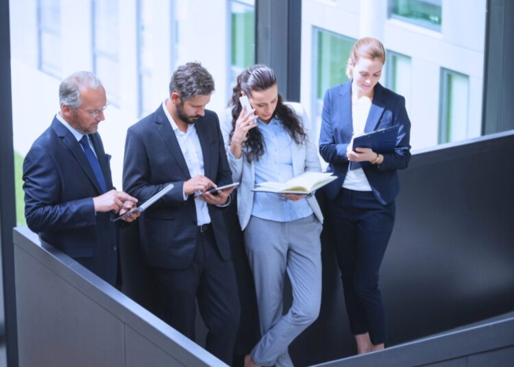 Grupo de profissionais em um ambiente corporativo, utilizando dispositivos móveis e materiais de leitura, representando diferentes perfis financeiros e estratégias de negócios.